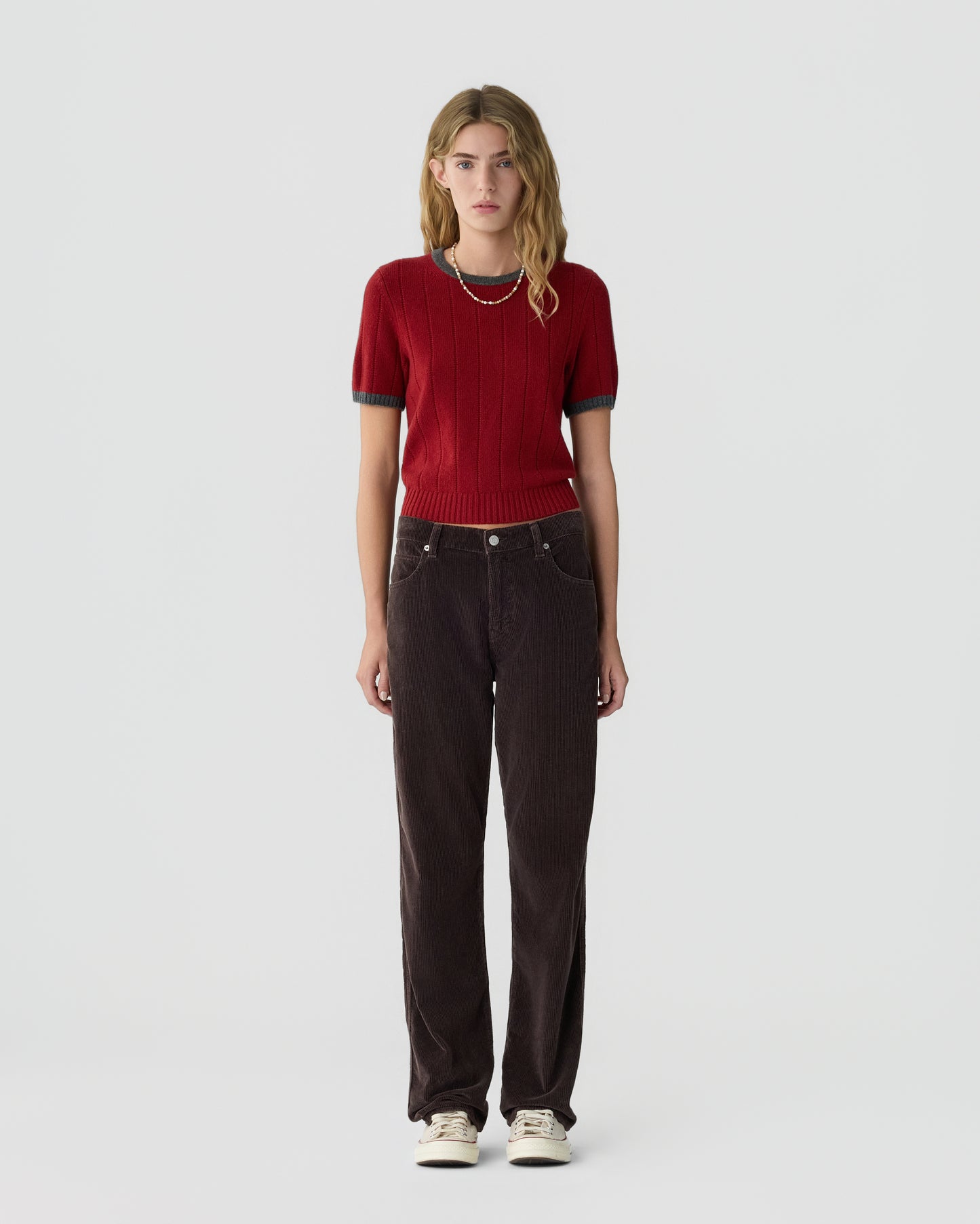Young woman with blonde wavy hair wearing a red short-sleeve cashmere sweater paired with dark brown wide-leg pants and white sneakers, standing against a plain white background.