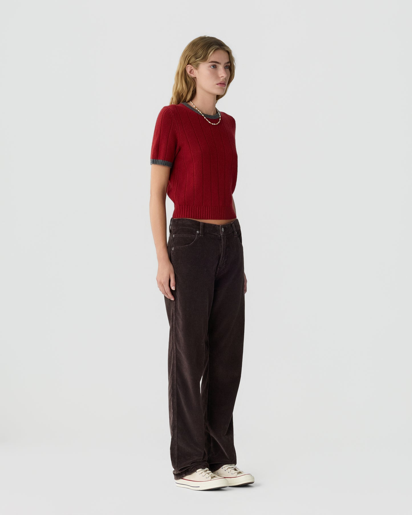 Young woman wearing a red short-sleeve knit top, dark brown wide-leg pants, and white sneakers standing against a plain light background.