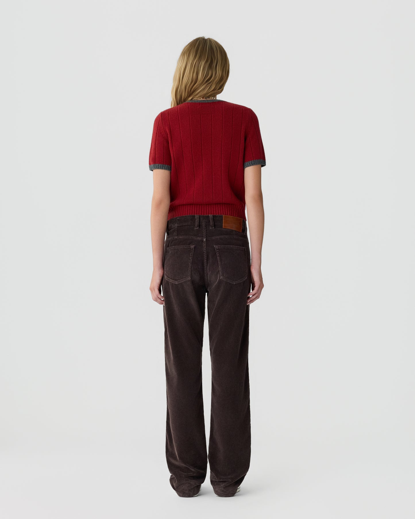 Woman seen from behind wearing a red short-sleeve knitted top and loose dark brown corduroy pants against a plain white background.