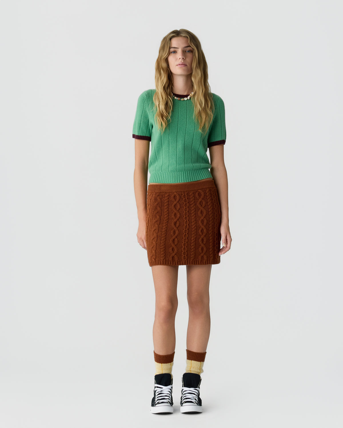 Woman wearing a green short-sleeve knit top paired with a brown cable-knit mini skirt, multi-colored socks, and black high-top sneakers standing against a plain white background.