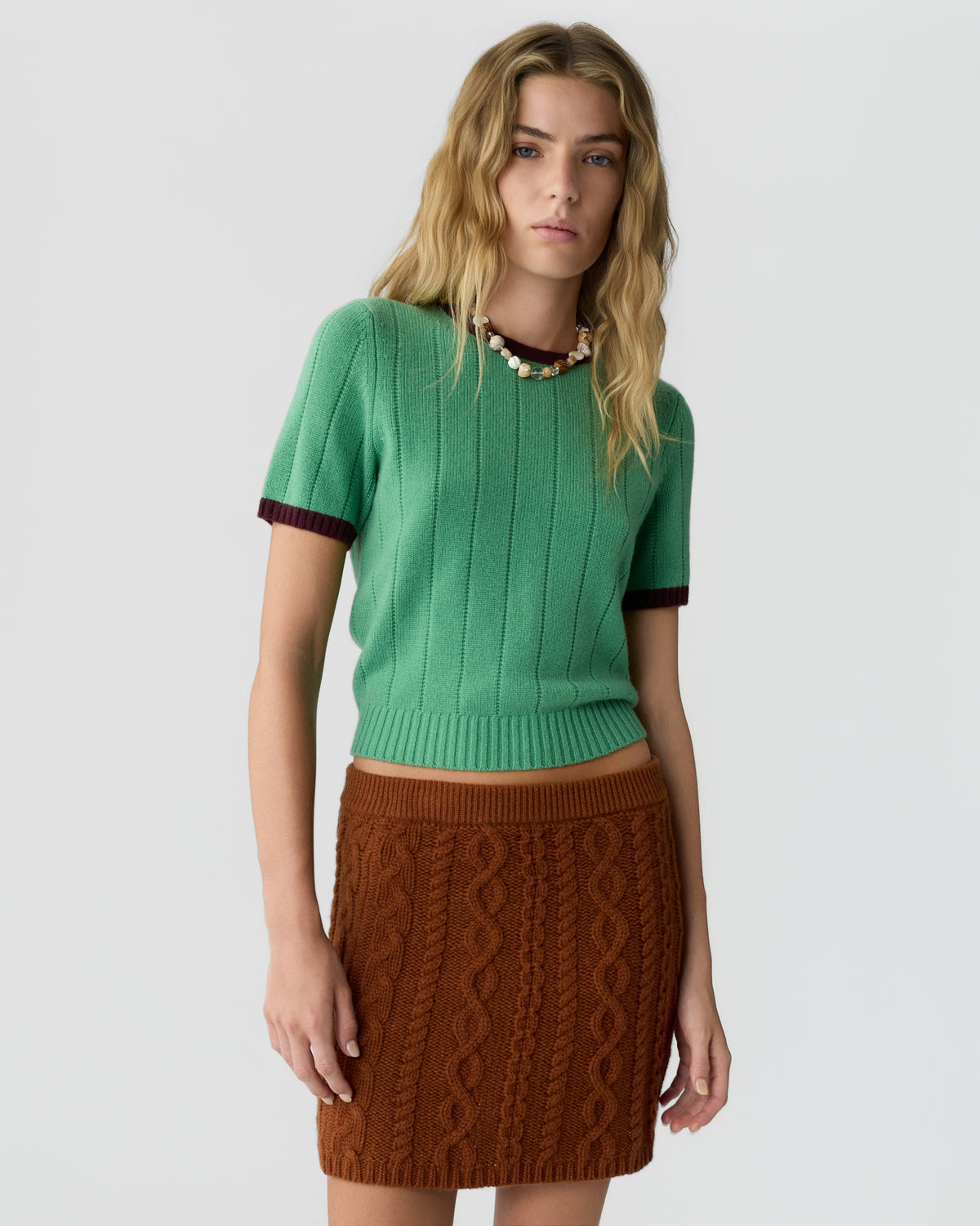 Young woman with wavy blonde hair wearing a green short-sleeve knit sweater with dark trim and a brown textured knit skirt against a plain light background.