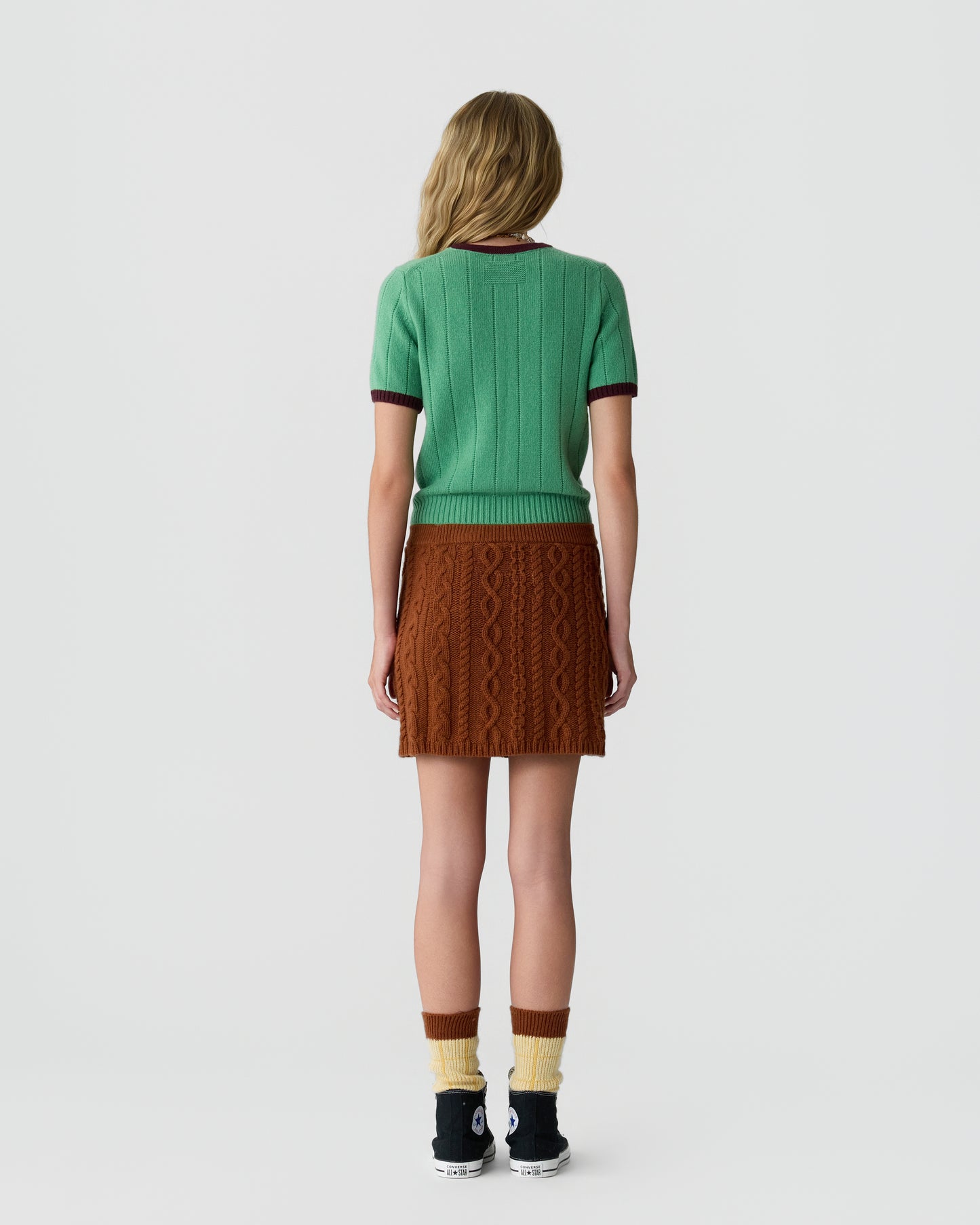 Woman wearing a green short-sleeve ribbed top with dark trim, a brown textured cable knit skirt, multi-colored socks, and black sneakers, standing facing away against a plain background.
