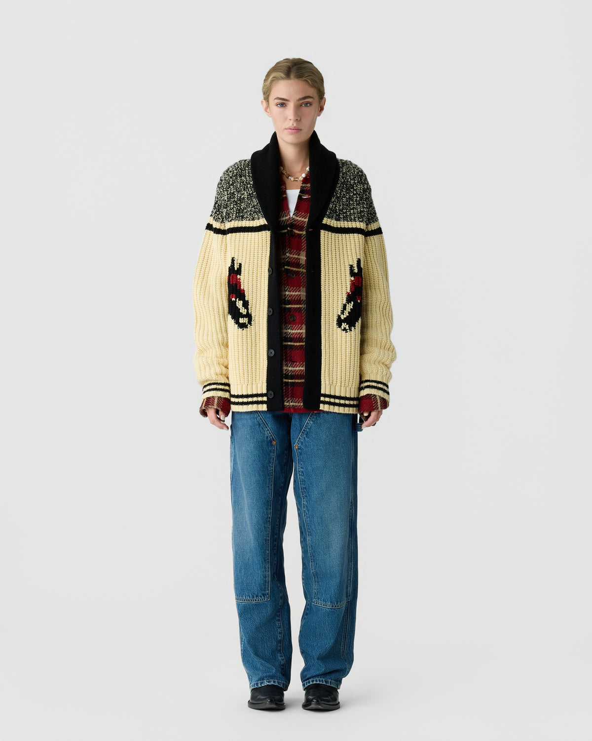 Full-length person wearing a patterned cream, black, and red cardigan with pocket designs, blue jeans, and black shoes against a light gray background