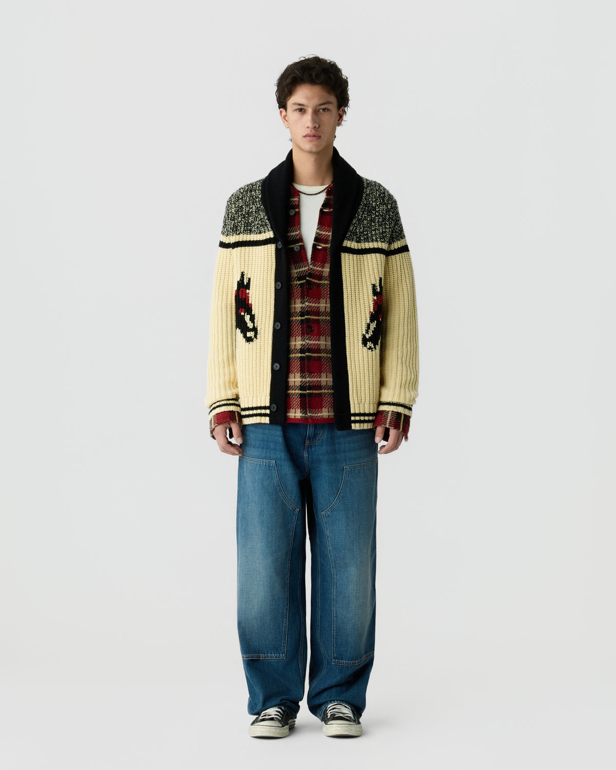 Full-length portrait of a man wearing a patterned cream and dark cardigan over a red plaid shirt, relaxed blue jeans, and casual sneakers against a light gray background