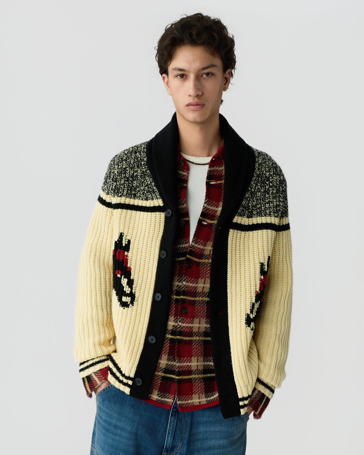 Young man wearing a cream and black patterned knit cardigan over a red plaid shirt with blue jeans against a light gray background
