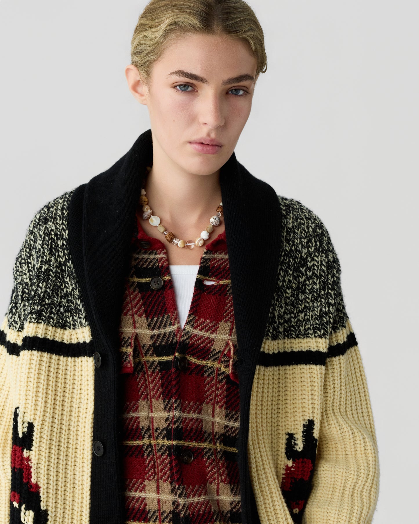 Model wearing a black cream and red patterned knit cardigan over a red plaid shirt with a beaded necklace against a light gray background