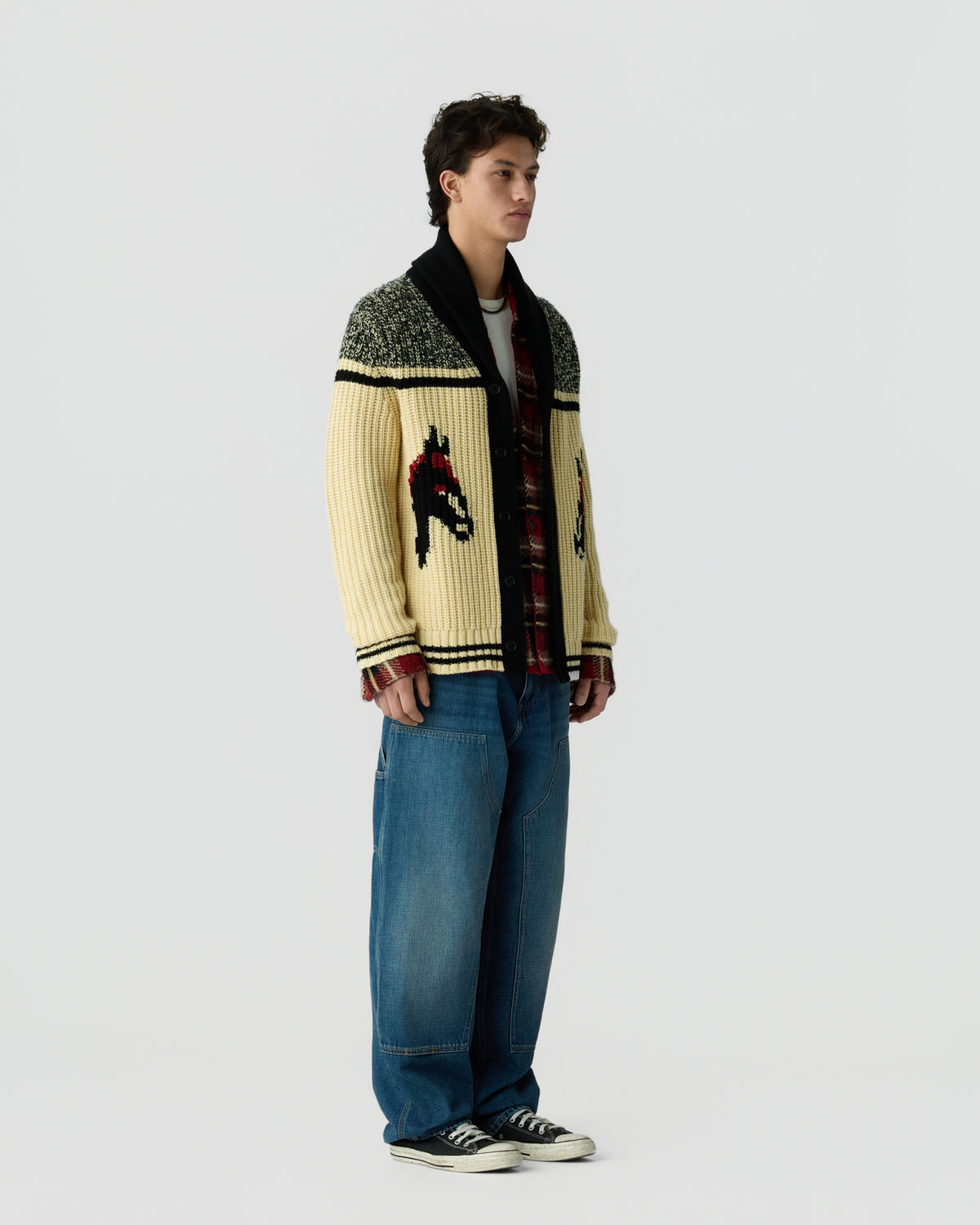 Full length view of a person wearing a patterned cream and black cardigan with graphic details blue jeans and black and white sneakers against a light gray background