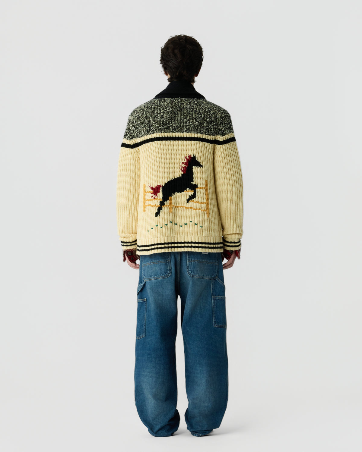 Back view of a person wearing a patterned cream and green knit sweater with a horse graphic and loose blue jeans against a plain light background