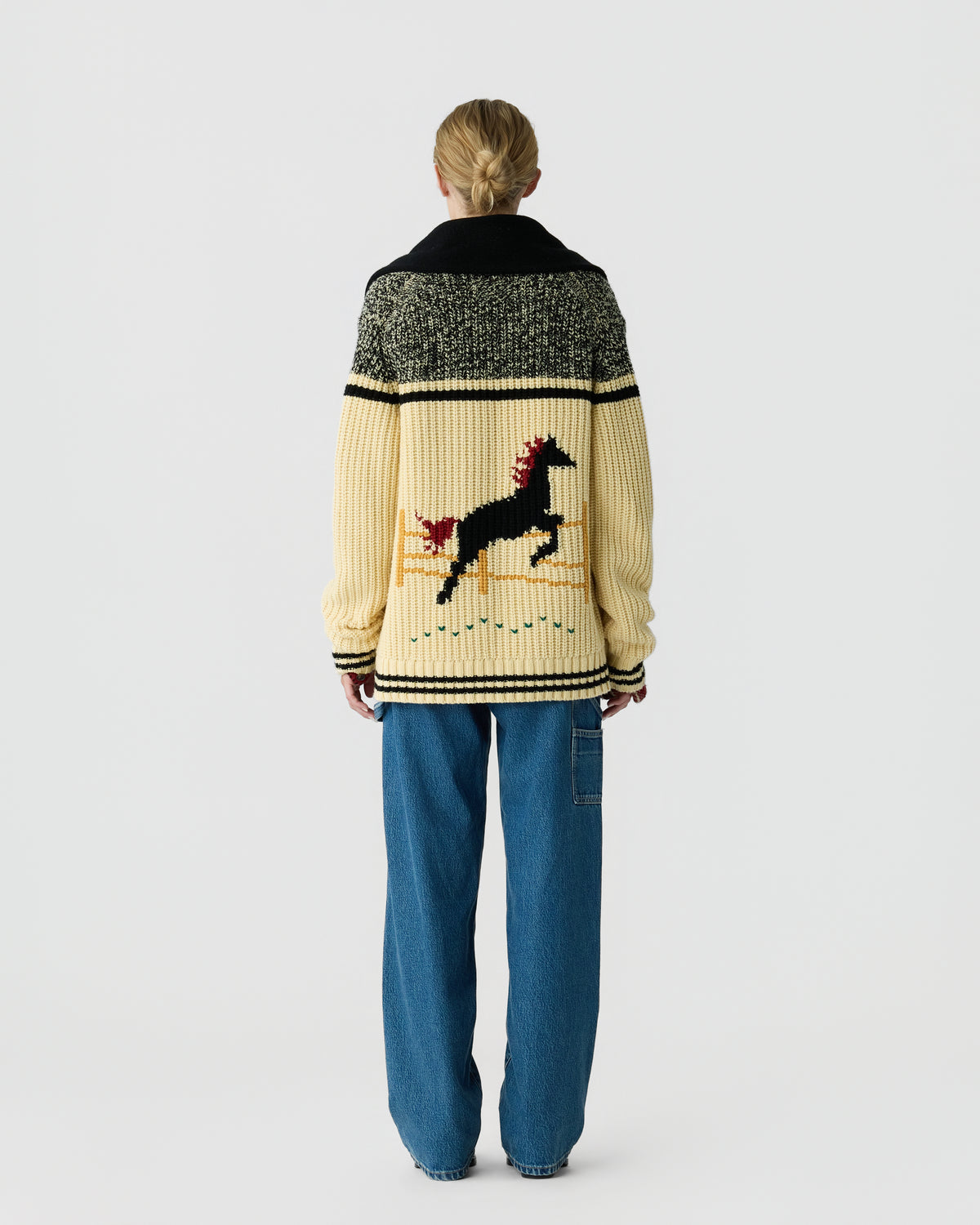 Person viewed from behind wearing a cream and dark green patterned cardigan with a reindeer motif, blue jeans, and hair tied back against a light gray background