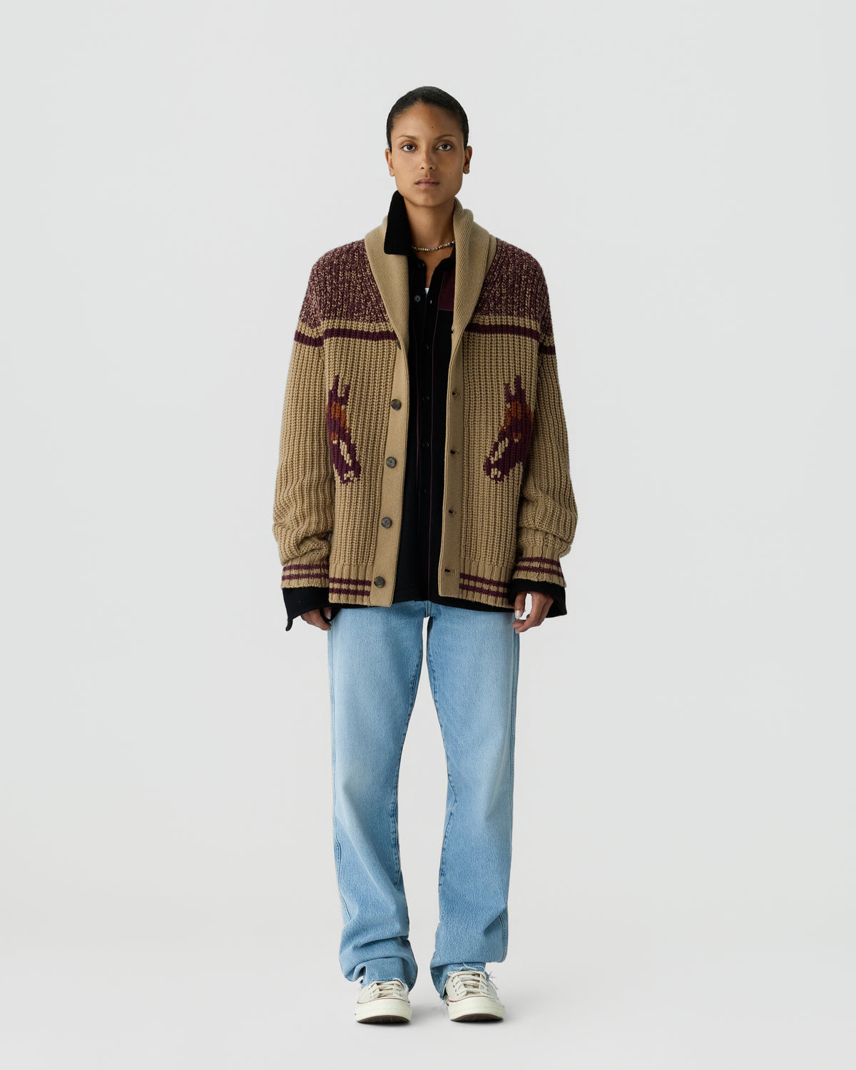 A person wearing a beige and brown knitted cardigan with a horse design on the front over a black shirt, paired with light blue jeans and white sneakers, standing against a plain white background.