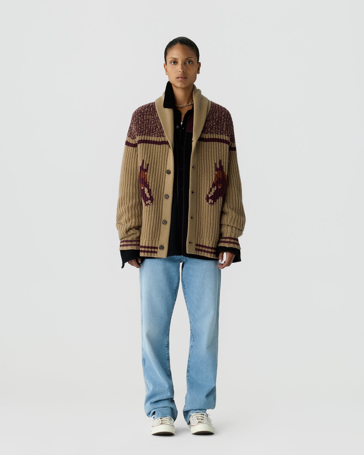 A person wearing a beige and brown knitted cardigan with a horse design on the front over a black shirt, paired with light blue jeans and white sneakers, standing against a plain white background.