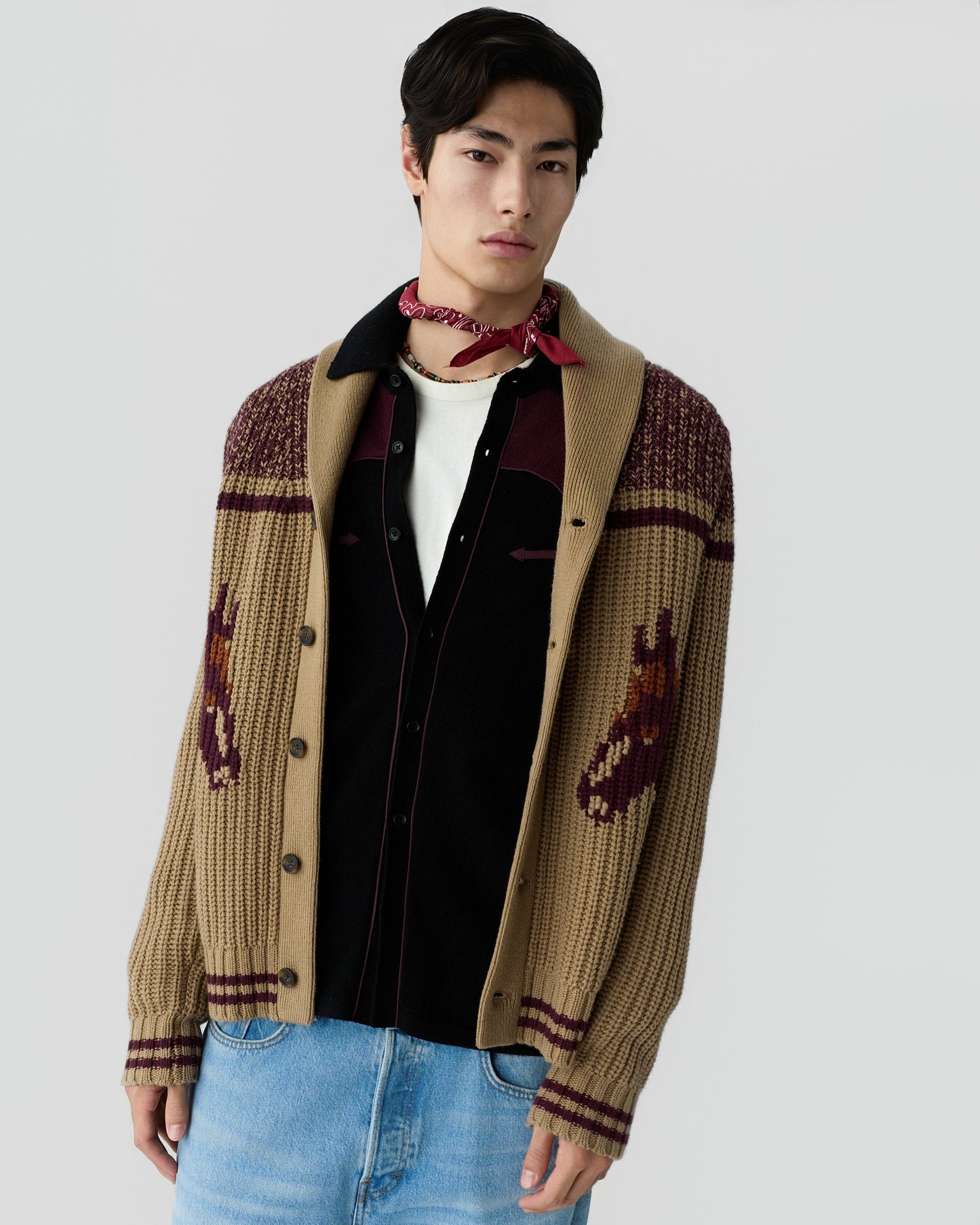 Young man wearing a beige and maroon knit cardigan with a horse pattern, a black and burgundy shirt, light blue jeans, and a red bandana tied around his neck against a plain background.