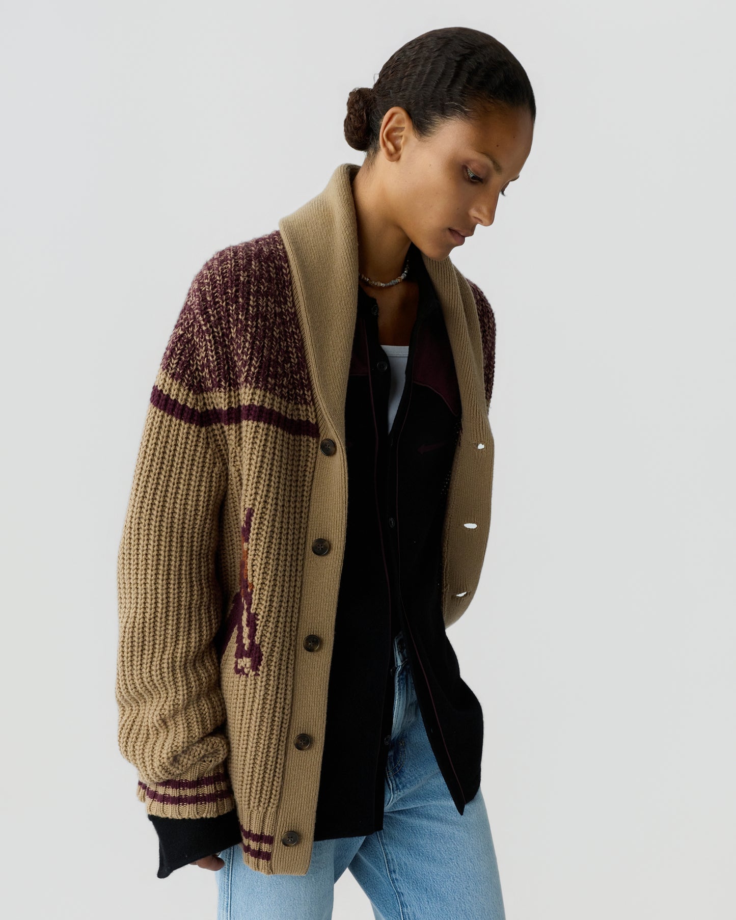 Woman with short dark hair styled in a bun wearing a chunky knit cardigan with brown and maroon patterns over a black jacket and light blue jeans against a neutral gray background