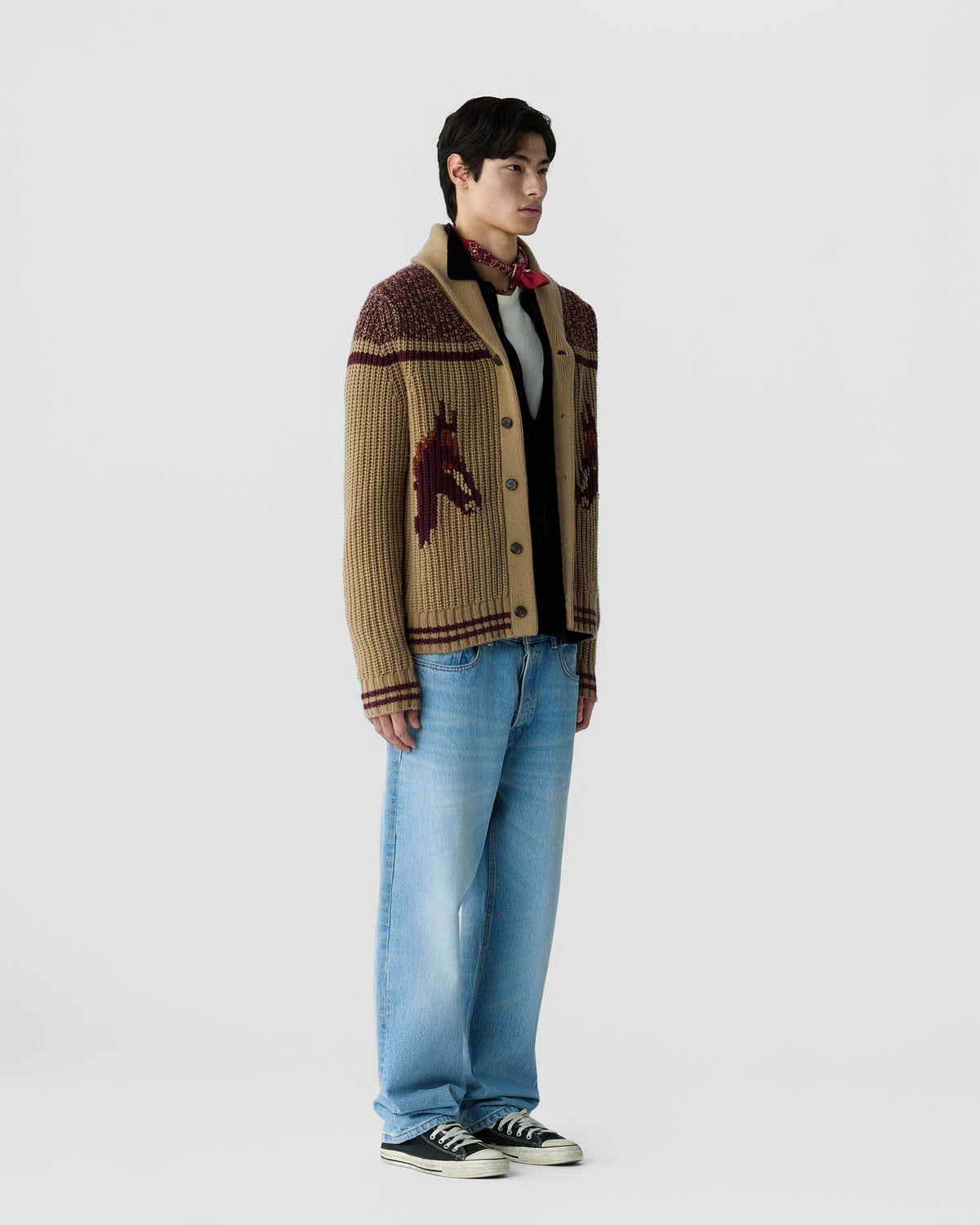 Man wearing a beige and brown knitted cardigan with horse designs on the sides, a light gray shirt underneath, light blue jeans, black sneakers, and a red patterned scarf tied around the neck.