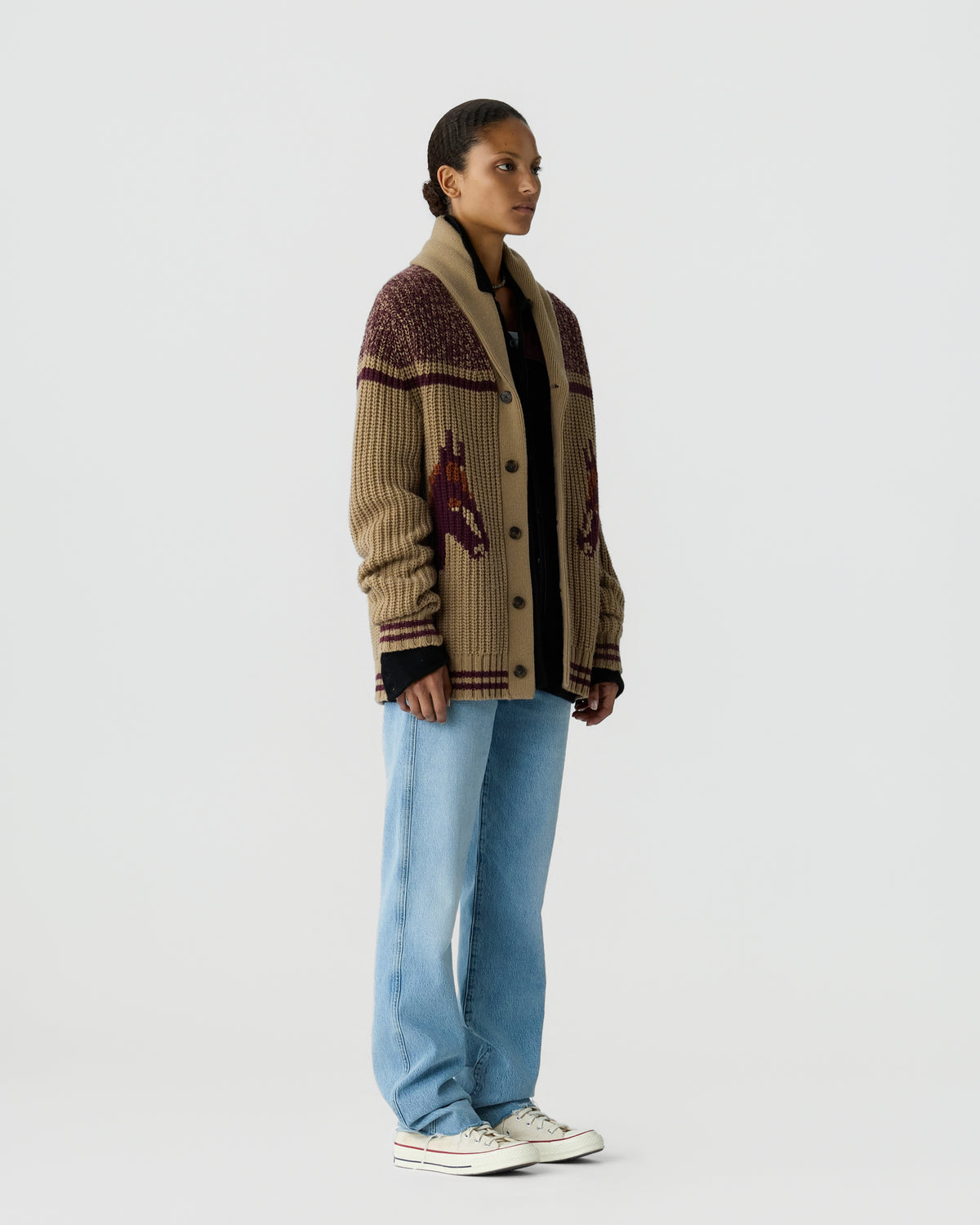 Woman standing against a plain background wearing a beige knit cardigan with a horse pattern, light blue jeans, a black top underneath, and white sneakers.