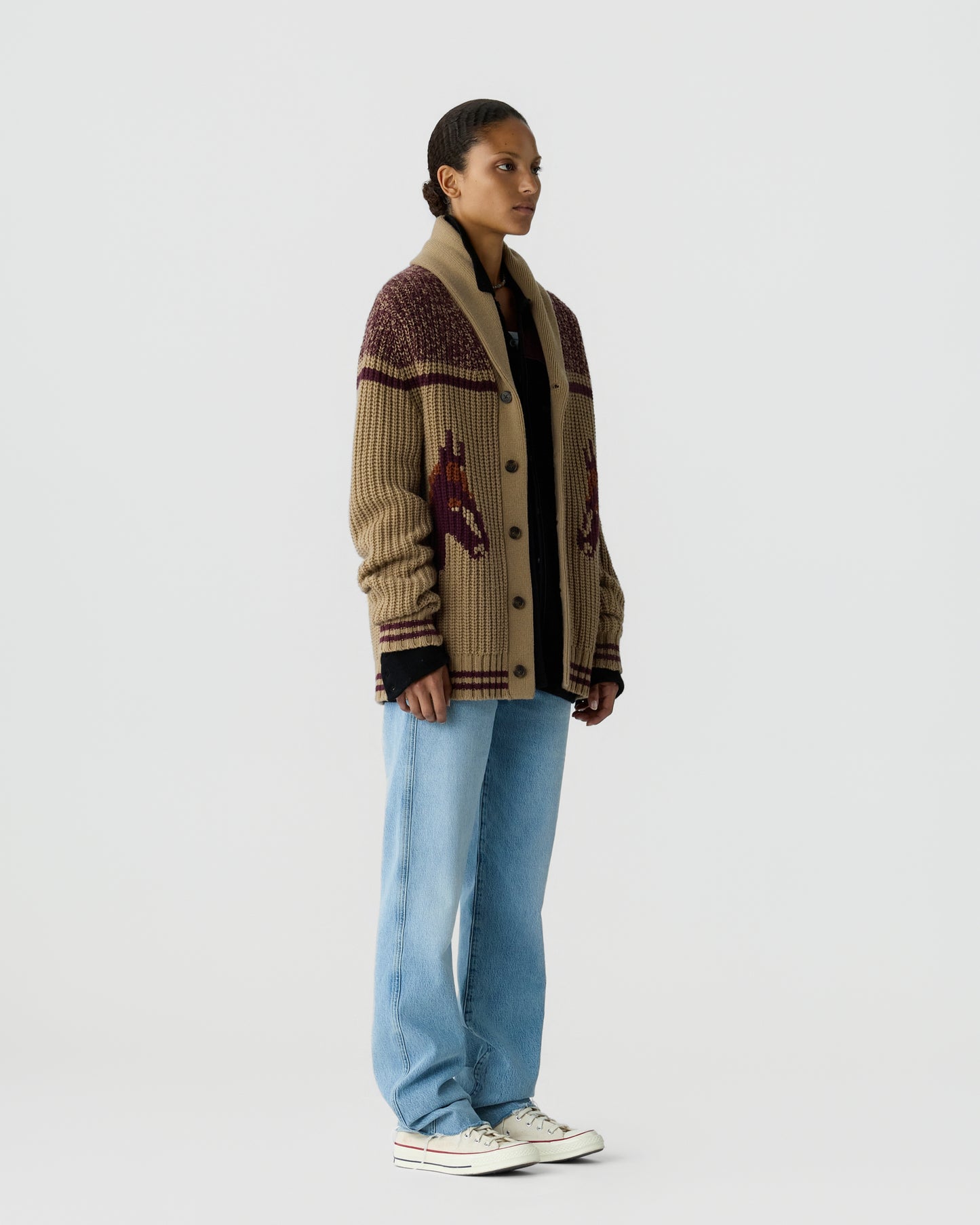 Woman standing against a plain background wearing a beige knit cardigan with a horse pattern, light blue jeans, a black top underneath, and white sneakers.