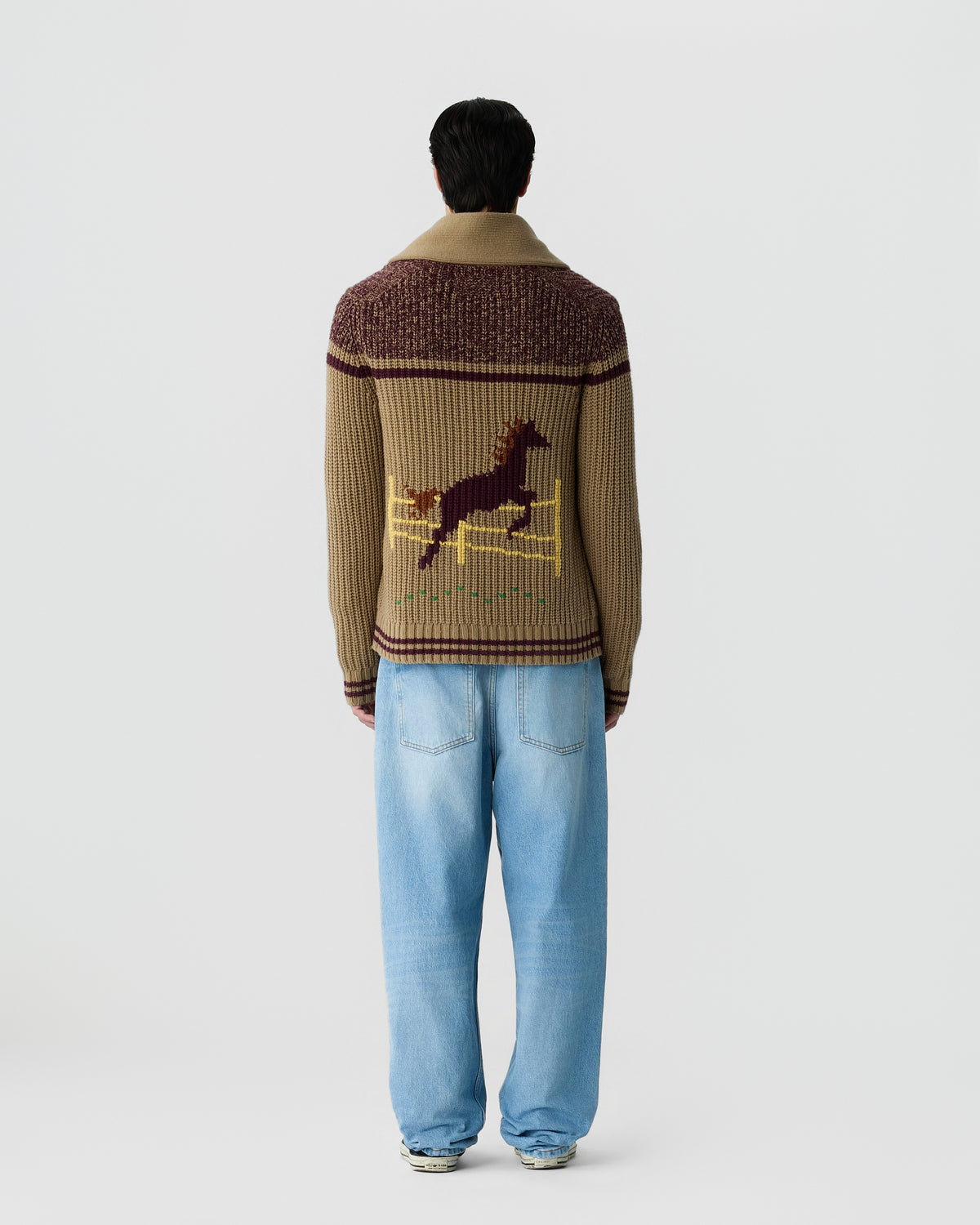 Man wearing a brown knit sweater with a horse jumping design on the back and light blue jeans, standing against a plain white background.
