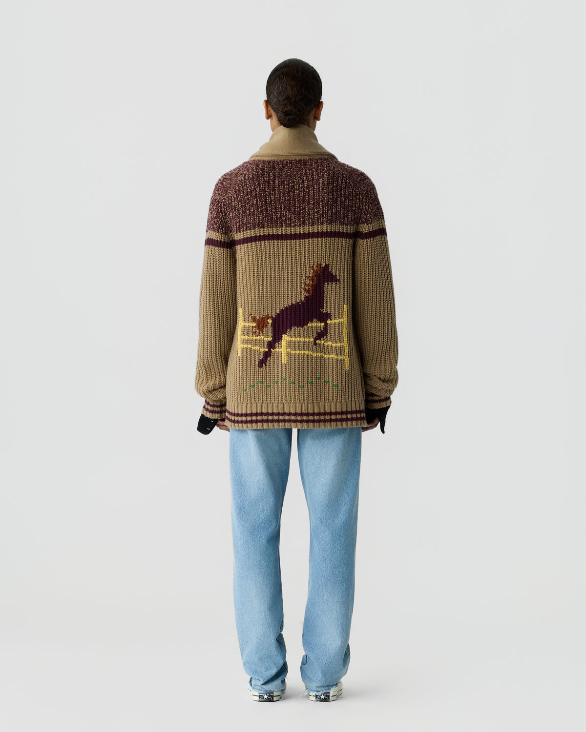 Person seen from behind wearing a chunky knit sweater with a horse jumping over a fence pattern, paired with light blue jeans and gray sneakers.