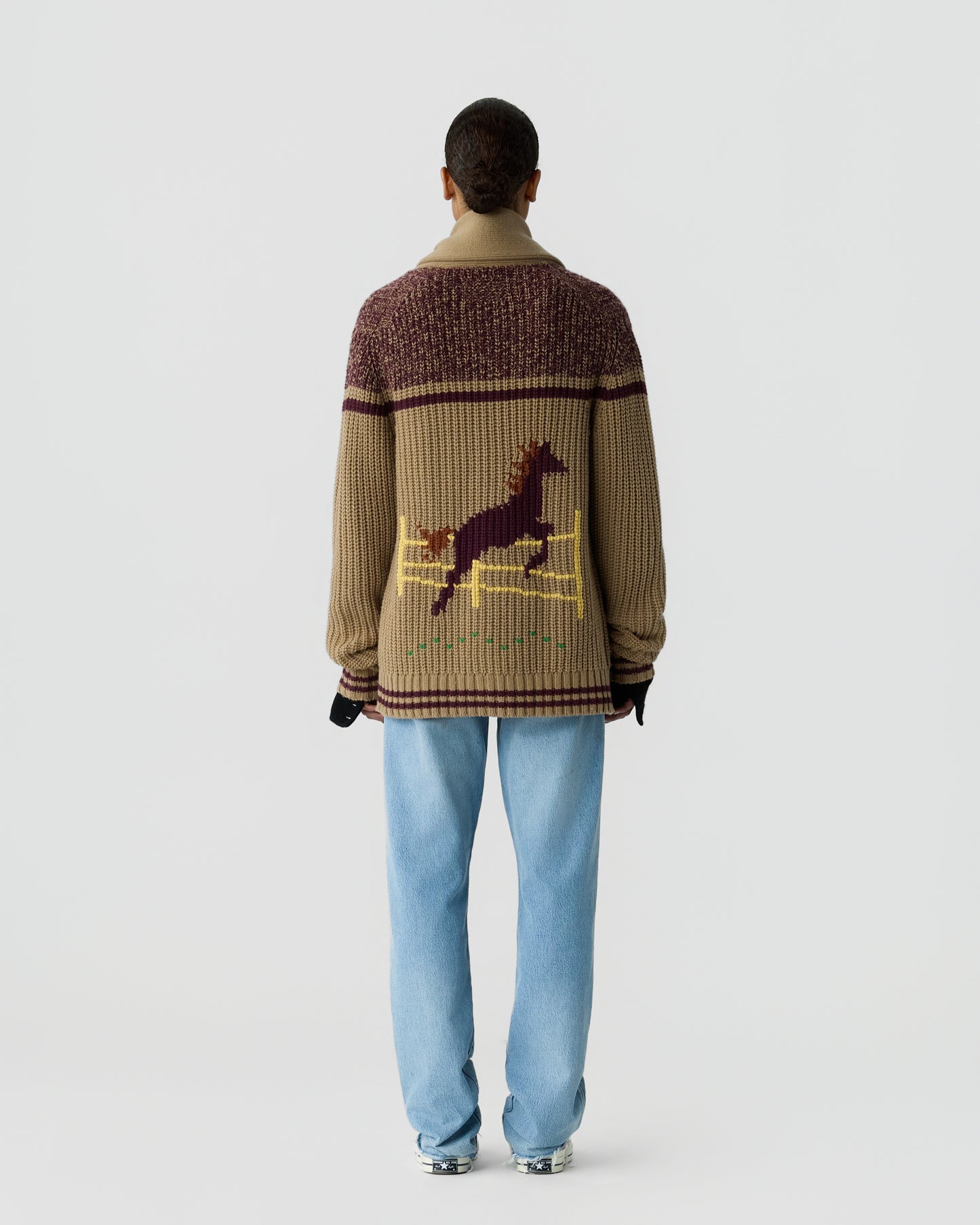 Person seen from behind wearing a chunky knit sweater with a horse jumping over a fence pattern, paired with light blue jeans and gray sneakers.