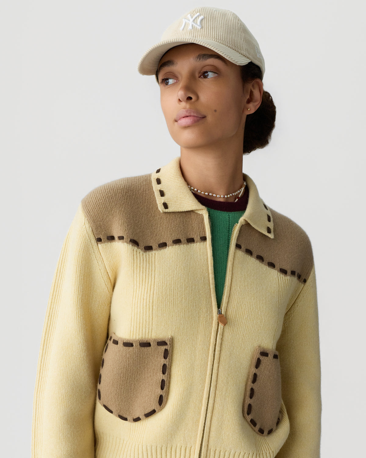 Woman wearing a beige and brown zip-up sweater with stitched pocket details and a light-colored baseball cap looking to the side against a plain background.