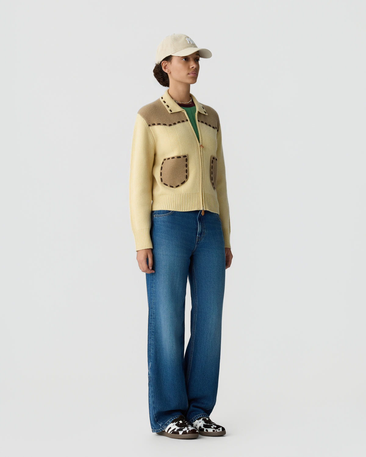 Woman wearing a cream and beige knitted cardigan with contrasting stitching, blue wide-leg jeans, a beige baseball cap, and black and white sneakers, standing against a plain white background.