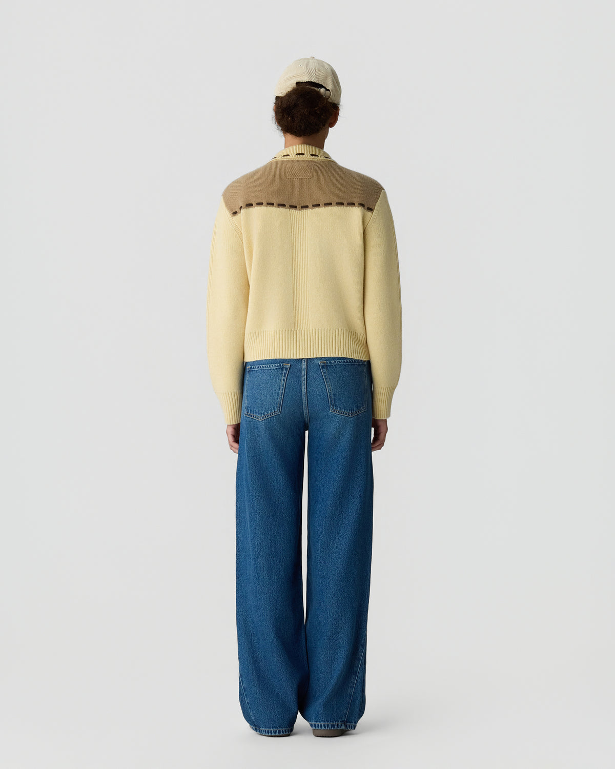 Person wearing a cream and tan color-blocked sweater with long sleeves and a beige cap, paired with high-waisted wide-leg blue jeans, standing facing away against a plain background.
