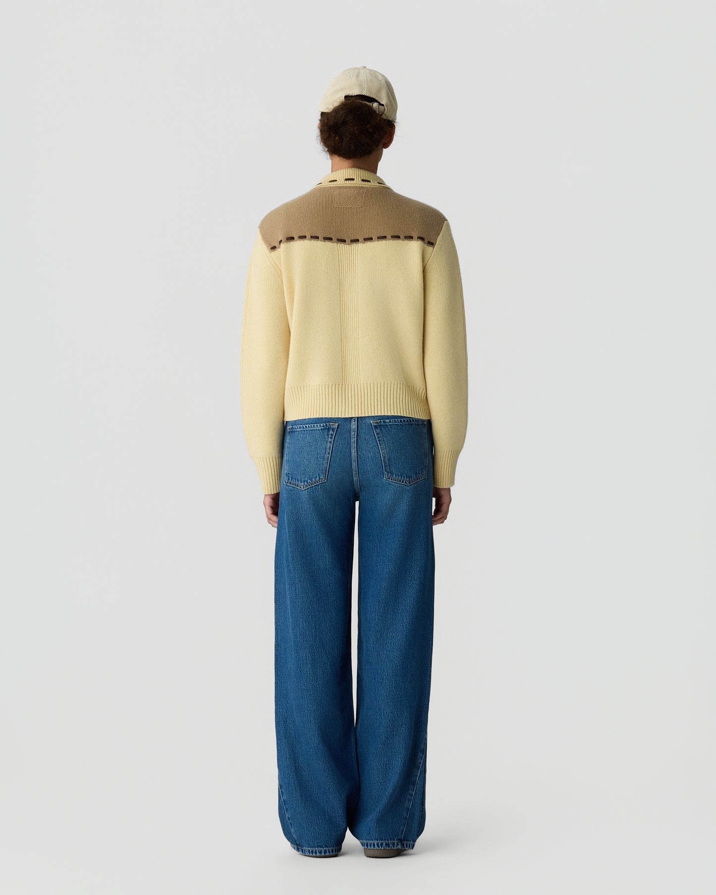 Person wearing a cream and tan color-blocked sweater with long sleeves and a beige cap, paired with high-waisted wide-leg blue jeans, standing facing away against a plain background.