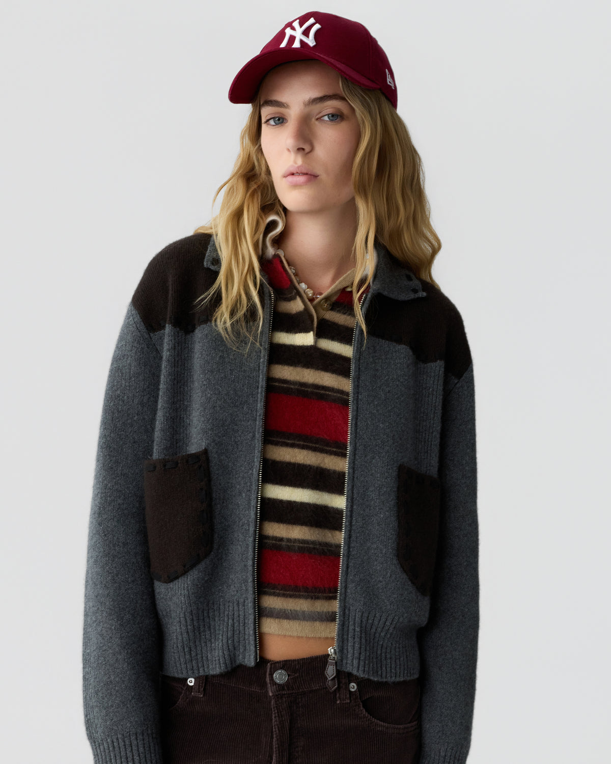 Young woman wearing a gray and dark brown zip-up cardigan over a striped sweater with red, beige, and brown tones, paired with dark brown pants and a maroon baseball cap.