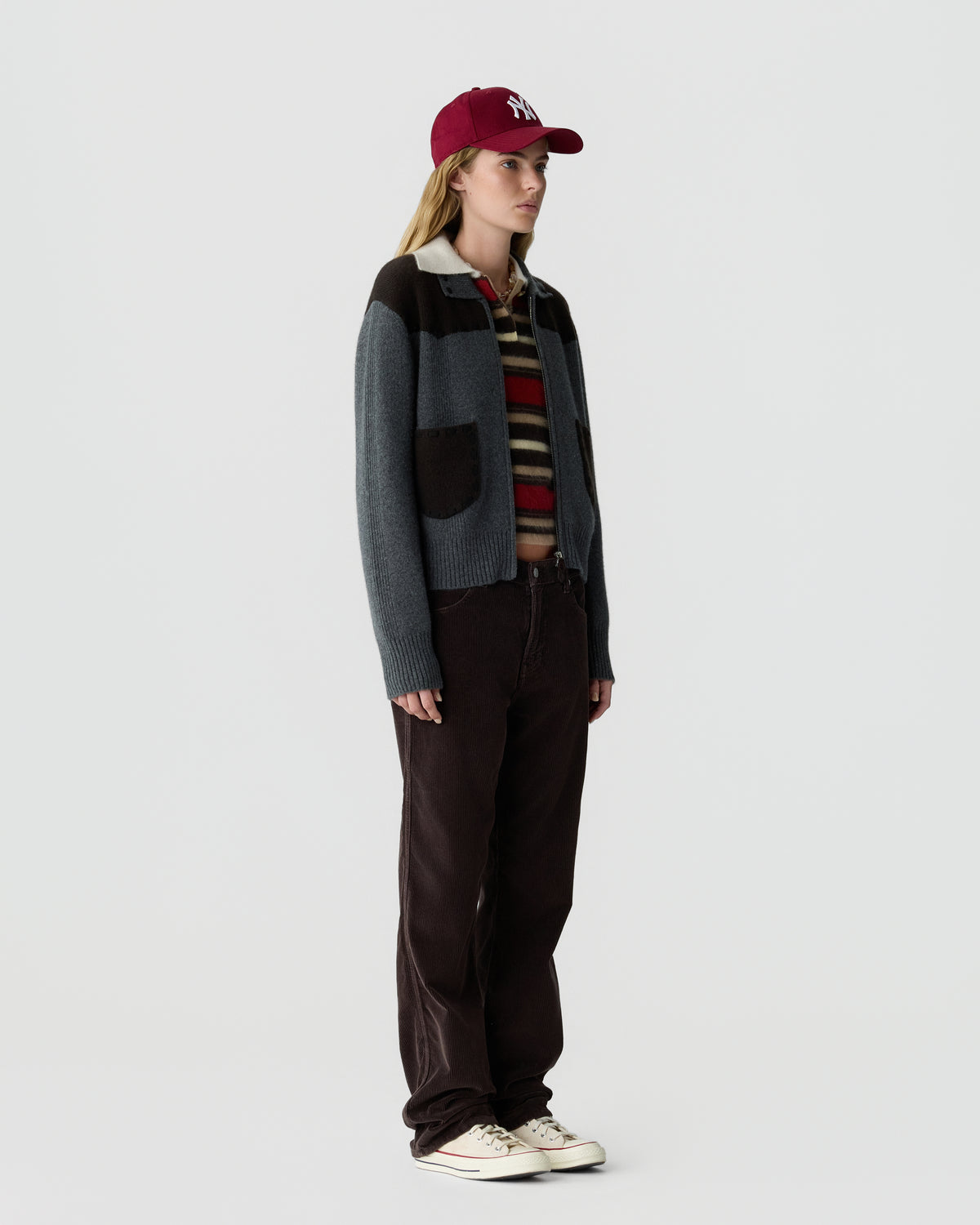 Woman wearing a burgundy cap, a striped cashmere sweater layered under a grey jacket with dark brown patches, dark pants, and white sneakers, standing against a plain background.
