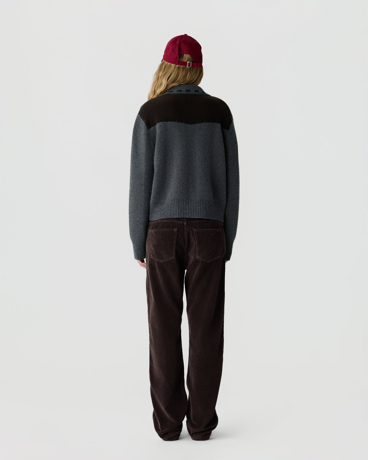 Person wearing a gray and dark brown sweater, brown pants, and a red cap, standing facing away against a plain light background.