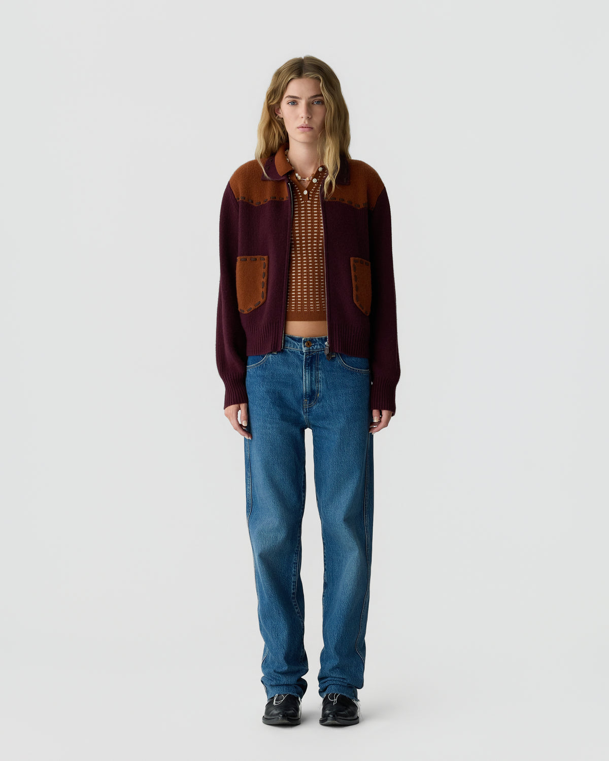 A person with long blonde hair wearing a maroon and brown knit cardigan over a patterned crop top, paired with high-waisted blue jeans and black shoes, standing against a plain light background.