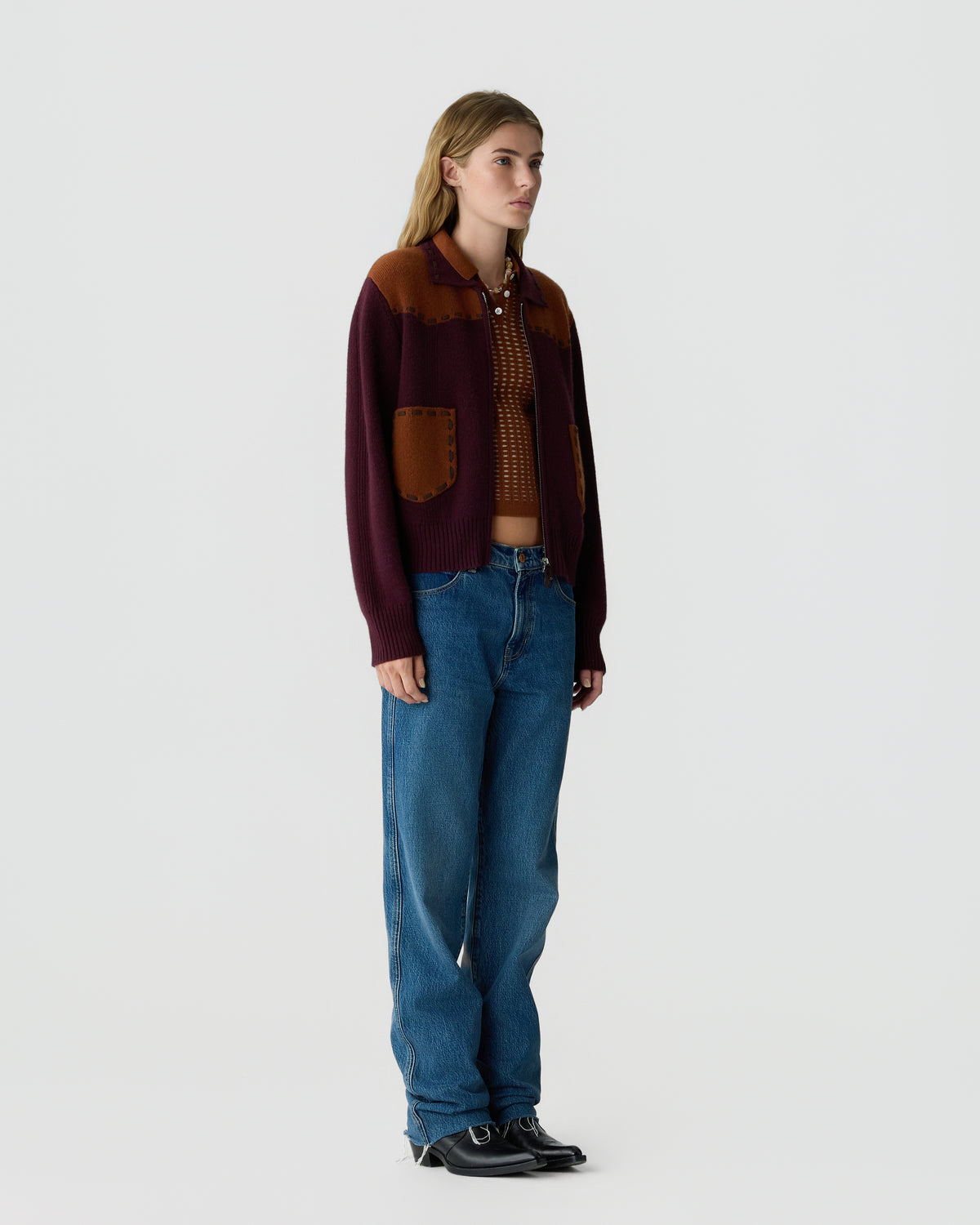 Woman standing wearing a burgundy and brown knitted cardigan over a brown patterned cropped top, blue jeans, and black pointed-toe shoes against a plain light background