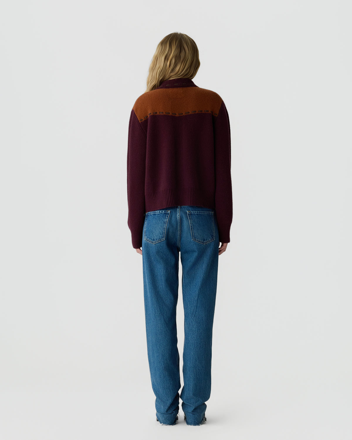 A person standing facing away wearing a color-blocked sweater with a rust and dark burgundy pattern, paired with blue denim jeans against a plain light gray background.