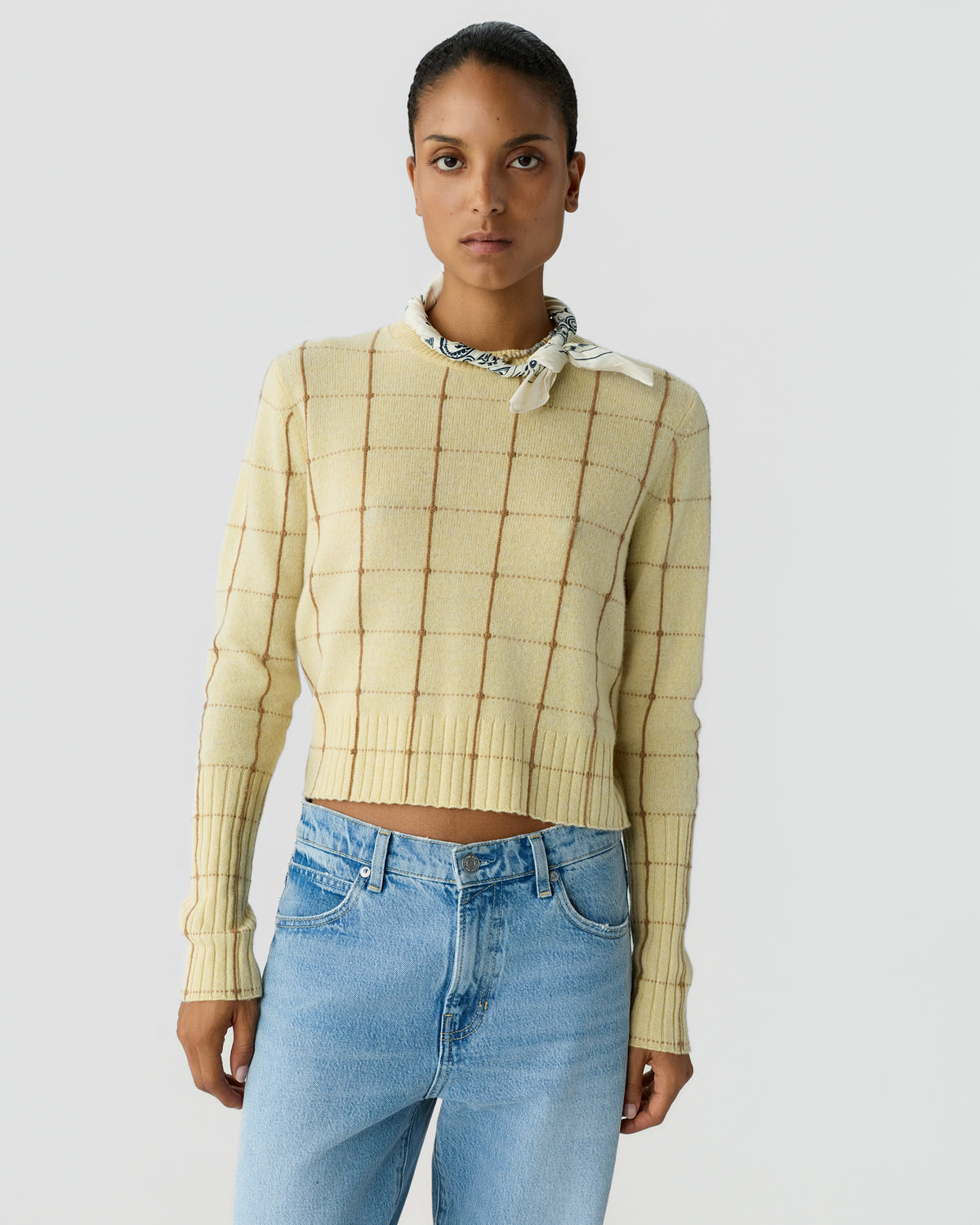 Woman wearing a cream-colored cashmere sweater with a subtle grid pattern, paired with light blue jeans and a patterned neck scarf against a plain background.