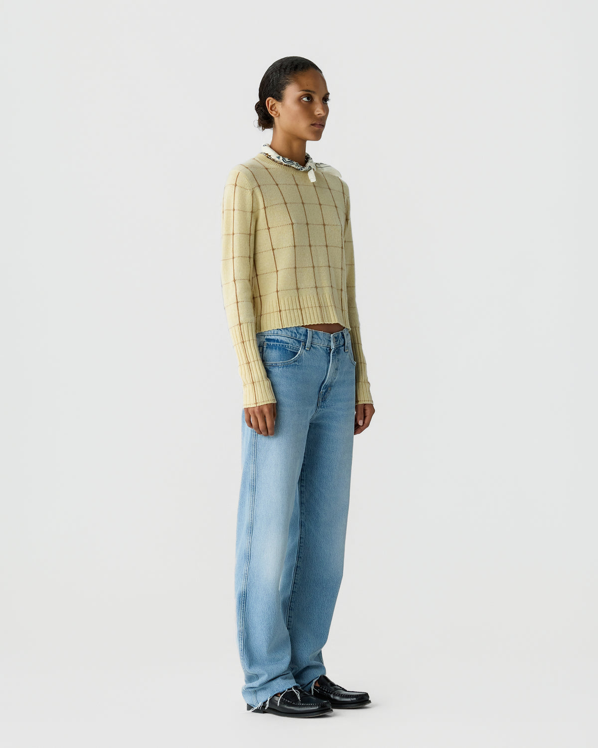 Woman wearing a beige long-sleeve cashmere sweater with a grid pattern, light blue wide-leg jeans, black loafers, and a white scarf tied around her neck against a plain white background.