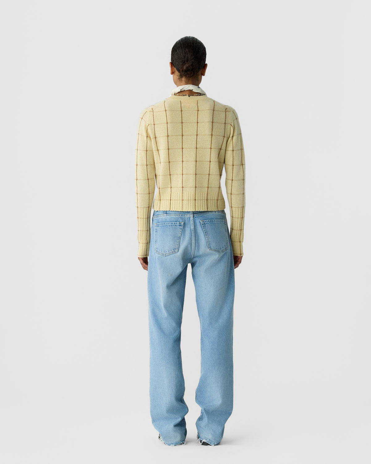 Person standing facing away wearing a cream-colored checkered cashmere sweater and light blue straight-leg jeans against a plain white background.