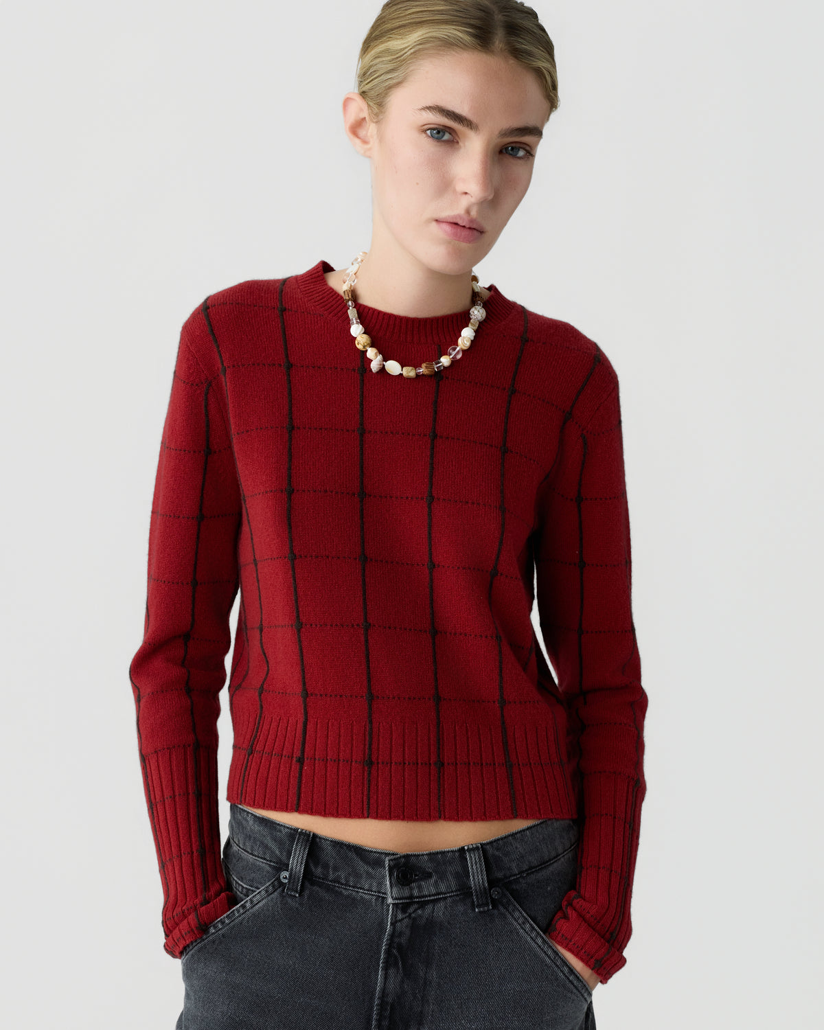 Model wearing a red checkered cashmere sweater with ribbed cuffs and hem paired with dark jeans and a layered pearl necklace