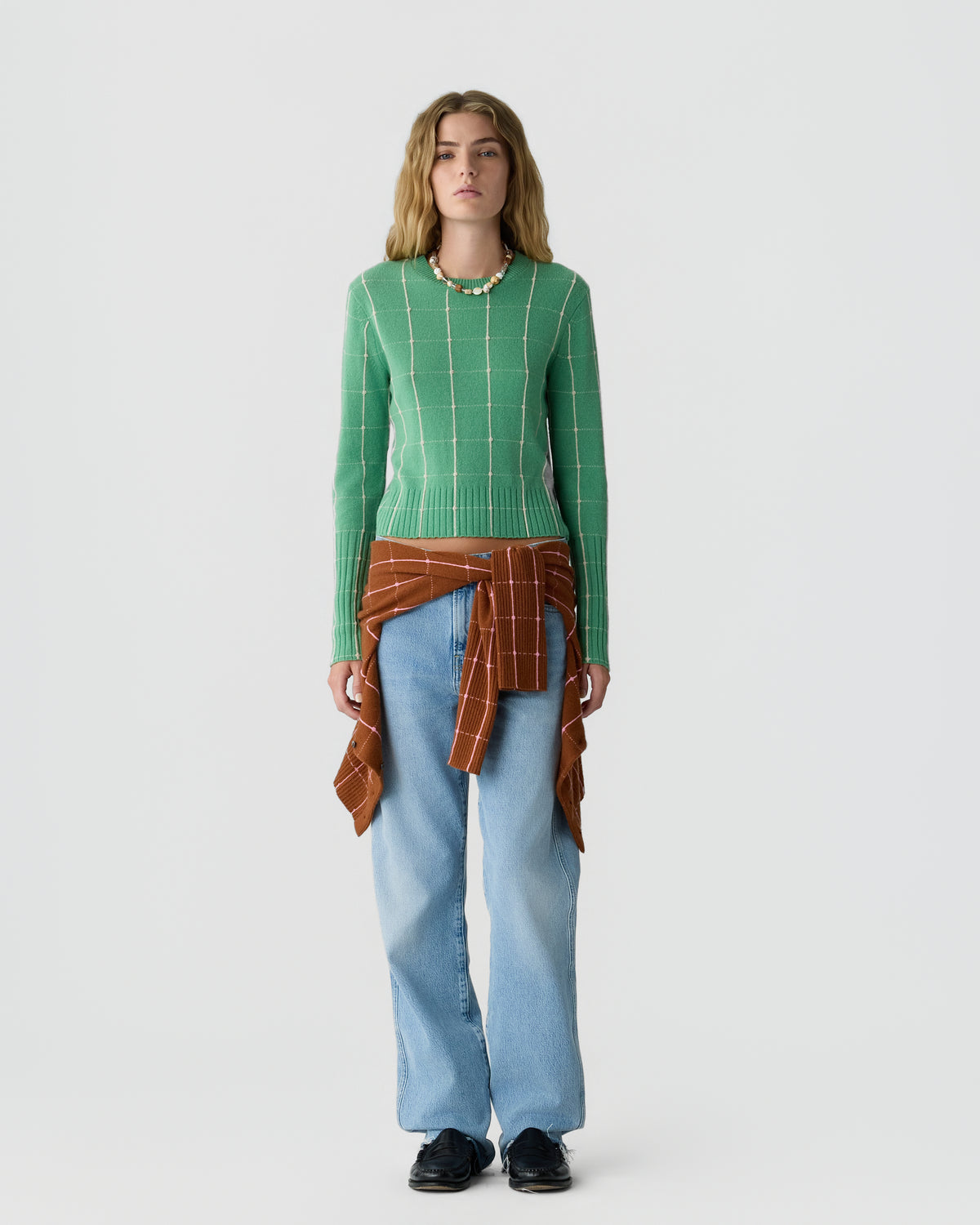 Woman standing against a plain background wearing a green long-sleeve sweater with a white grid pattern, light blue wide-leg jeans, black shoes, and a rust-colored shirt tied around her waist.