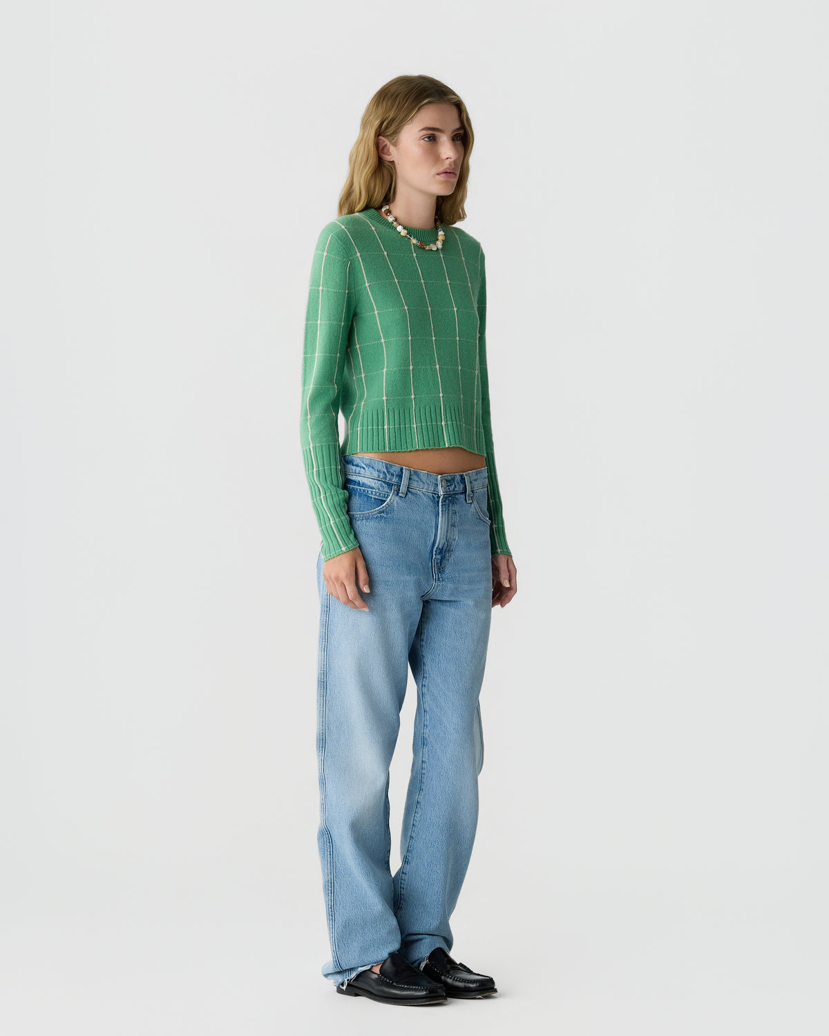 Woman wearing a green long-sleeve knit sweater with a white grid pattern, light blue loose-fitting jeans, black shoes, and a beaded necklace, standing against a plain background.