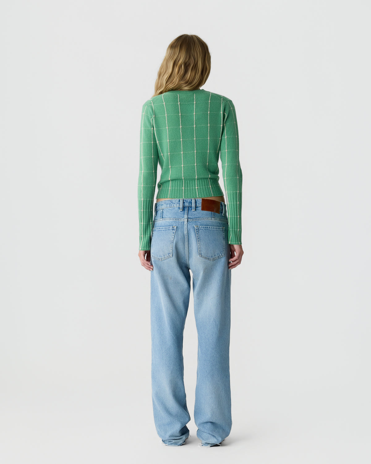 Woman seen from the back wearing a green long-sleeve sweater with a white grid pattern and light blue loose-fitting jeans against a plain white background.
