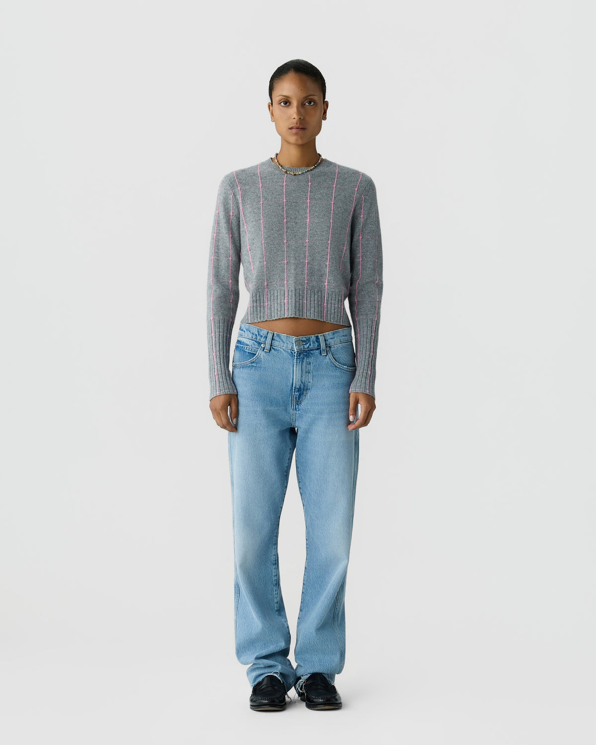 Person standing against a light background, wearing a gray striped cropped sweater, light blue jeans, and black shoes.
