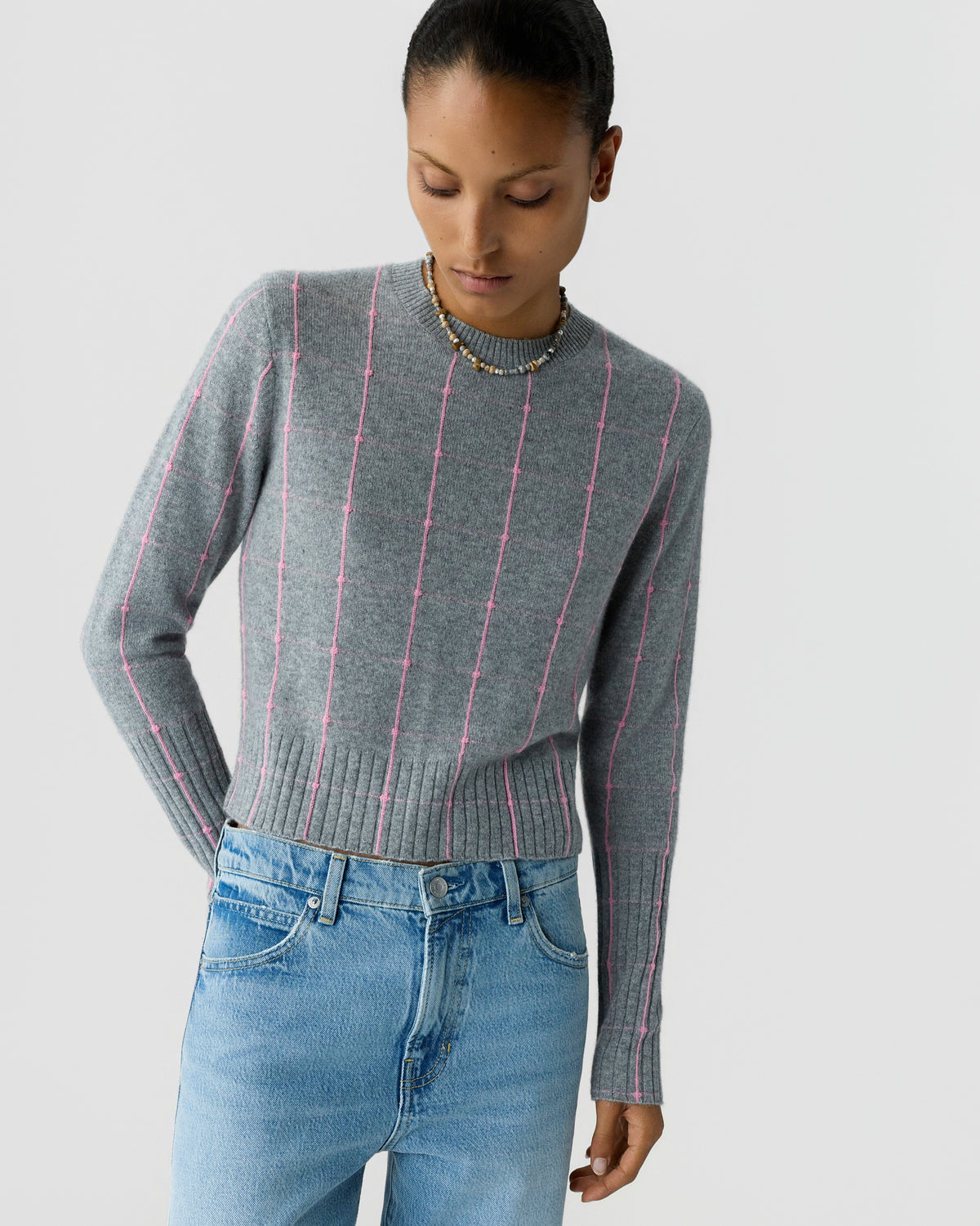 Woman wearing a gray knit sweater with pink vertical stripes and blue jeans while looking down against a light background