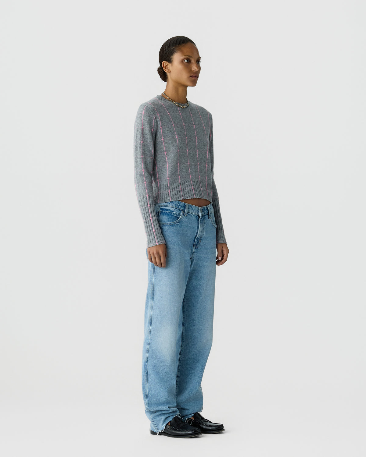 Standing model wearing a gray striped knit sweater with blue wide leg jeans and black shoes against a plain light gray background