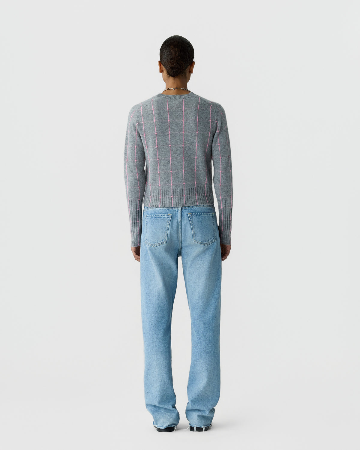Rear view of a person wearing a gray ribbed sweater with pink vertical stripes, light blue jeans, and black shoes against a plain light background.