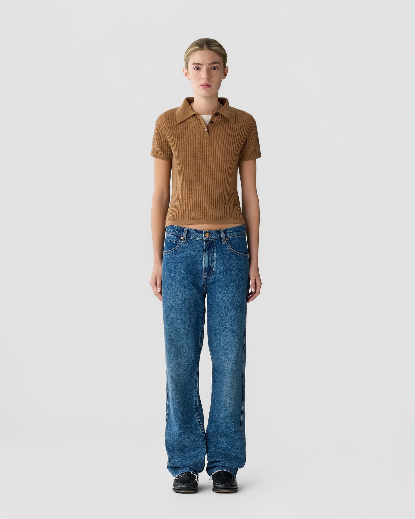 Model wearing a brown ribbed collared knit top with blue jeans and black shoes against a plain light gray studio background