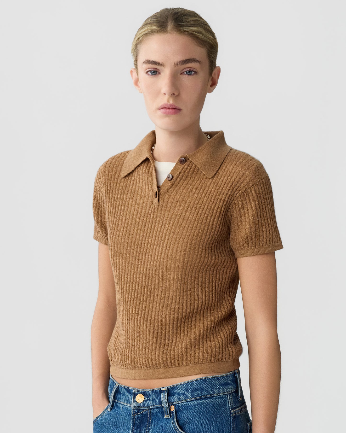Model wearing a brown ribbed cashmere polo shirt with a collared neckline and short sleeves, paired with blue jeans against a light gray background