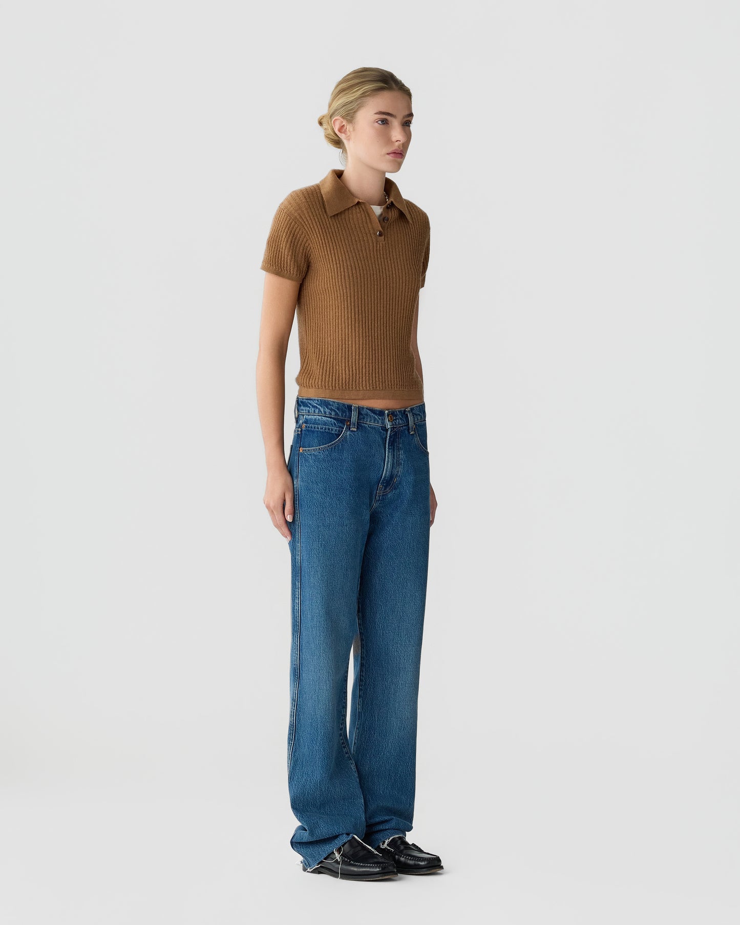 Model wearing a brown ribbed polo shirt with blue wide leg jeans and black shoes against a plain light gray studio background