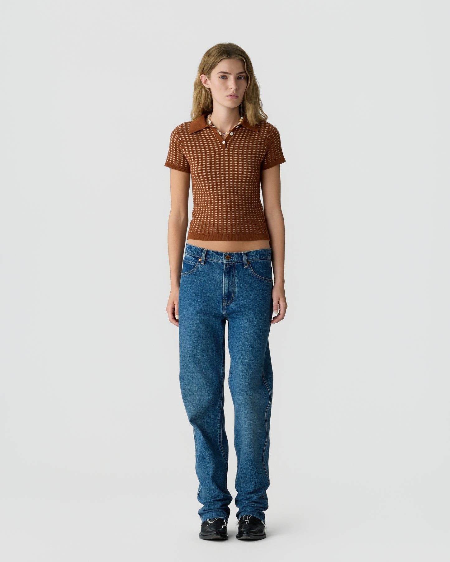 Woman wearing a short-sleeve brown patterned polo shirt paired with high-waisted blue jeans and black sneakers, standing against a plain light background.