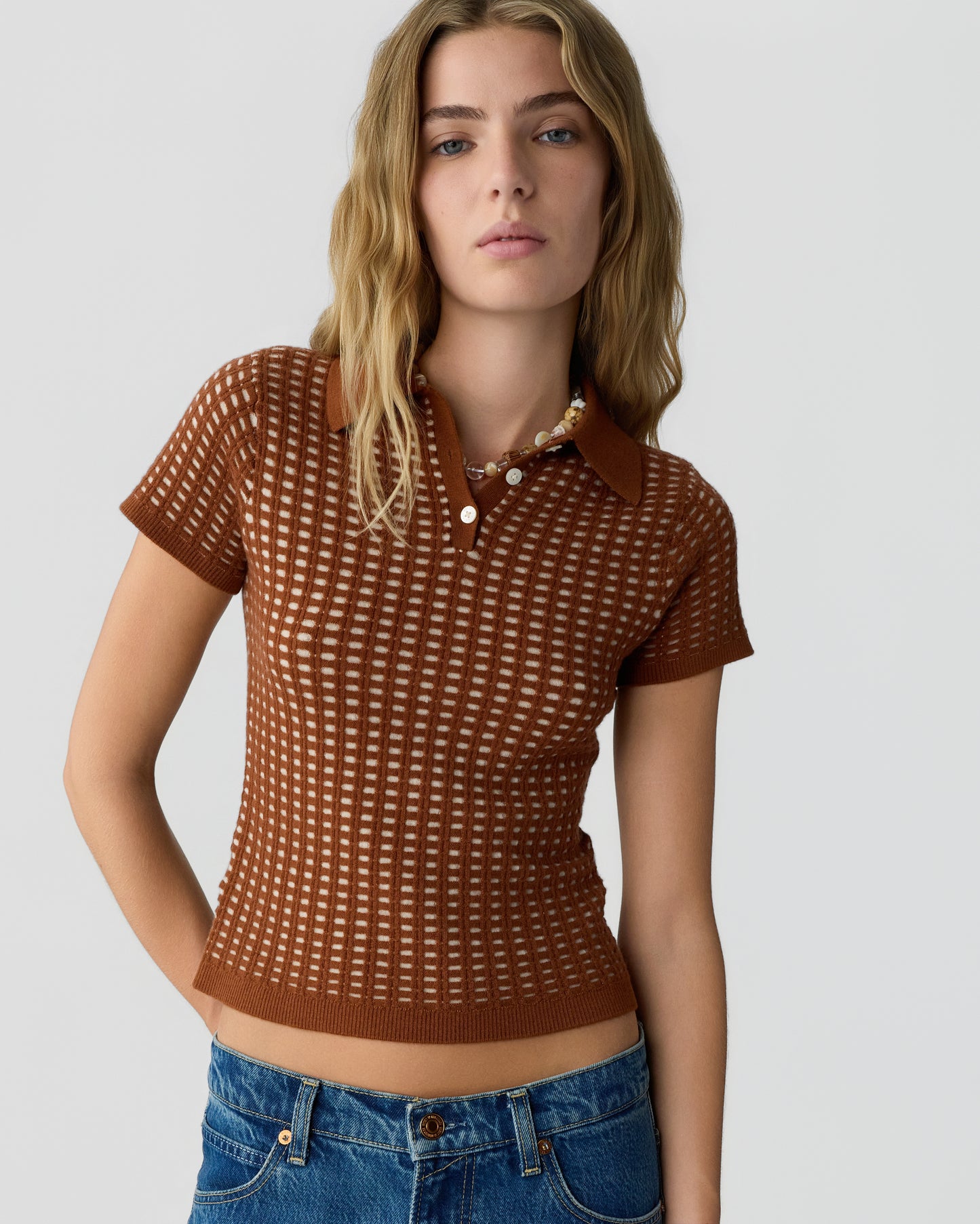 Young woman with long blonde hair wearing a short-sleeve brown knit polo shirt with a checkered pattern and blue jeans, standing against a plain light background.