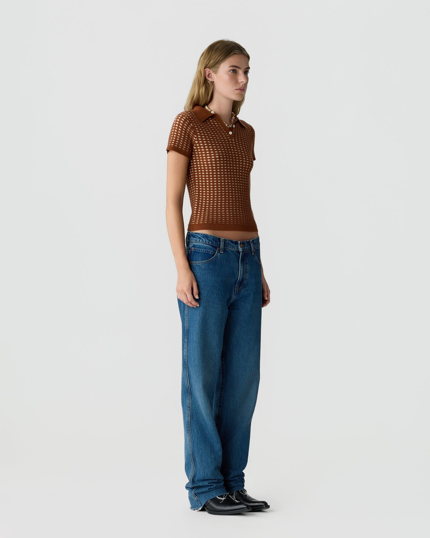 Young woman with long blonde hair wearing a brown short-sleeve knit polo shirt and loose blue jeans standing against a plain light gray background.